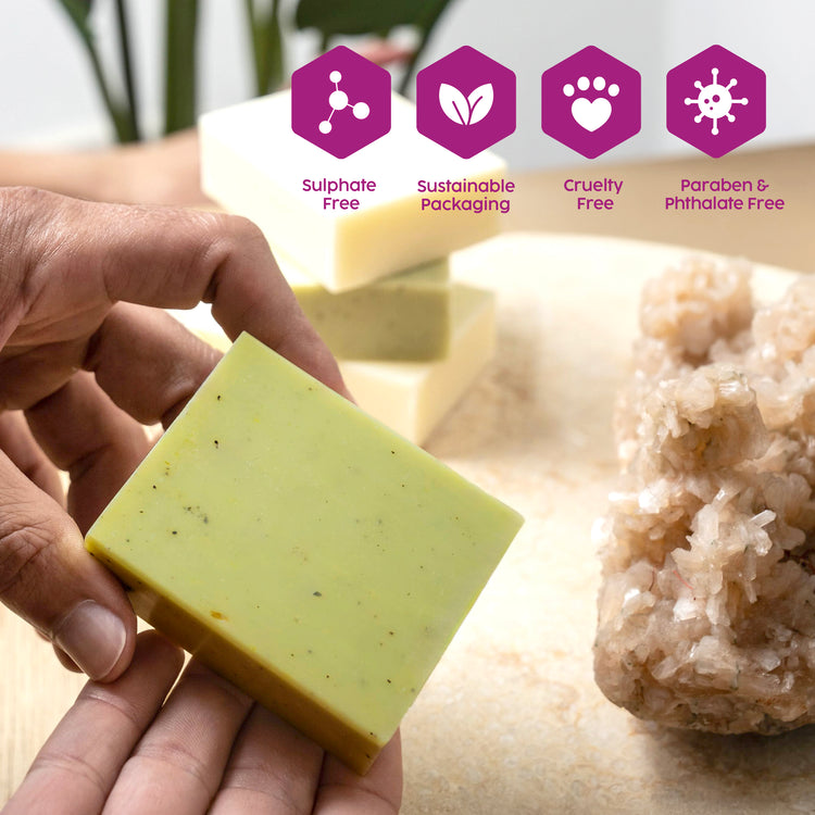 A hand holds a rectangular bar of pale green WashU3 soap with small dark specks and a crystal cluster in the background. Four purple icons at the top read 'Sulphate Free', 'Sustainable Packaging', 'Cruelty Free', and 'Paraben & Phthalate Free'.